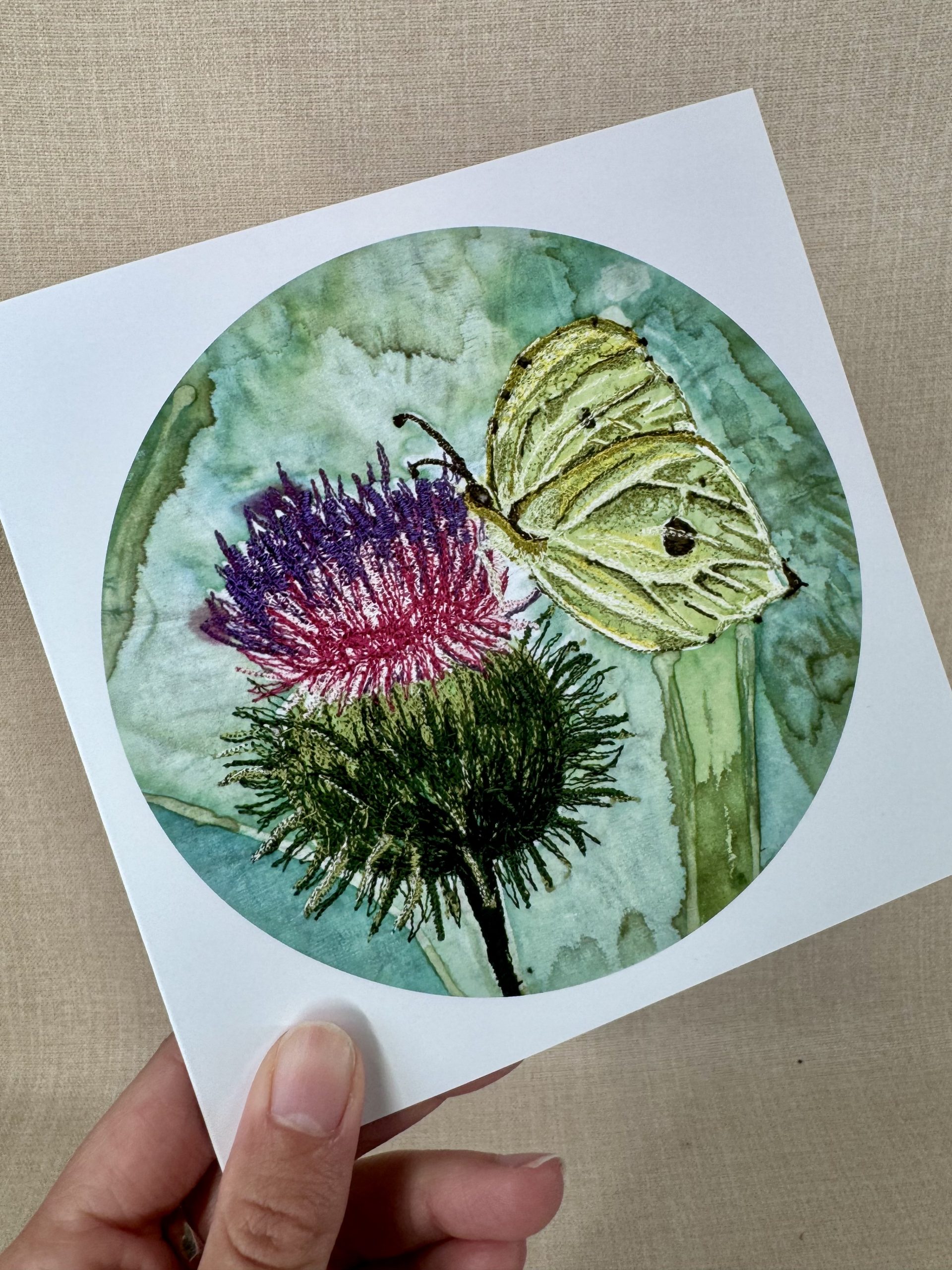 A hand holding a printed card featuring an original hand-painted embroidered Brimstone Butterfly artwork resting on a thistle.