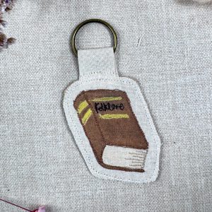 A photo of a Wild Minder Folklore book "Tiny Talisman" keyring. The book is brown with gold detail and black "Folklore" text on the cover.