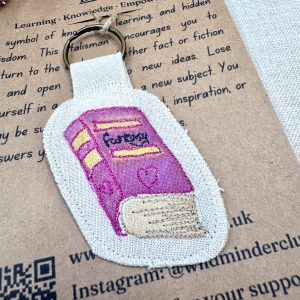 A close up angled photo of a Wild Minder fantasy book "Tiny Talisman" keyring displayed on it's backing card. The book is pink with gold detail and black "fantasy" text on the cover. The backing card is brown with black text.