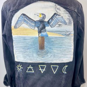 Wild Minder wearable art jacket featuring a hand-painted and free-motion embroidered cormorant drying its wings on a harbour post, with sea, cliffs, boat and symbolic sun, earth, water, air and moon embroidery.